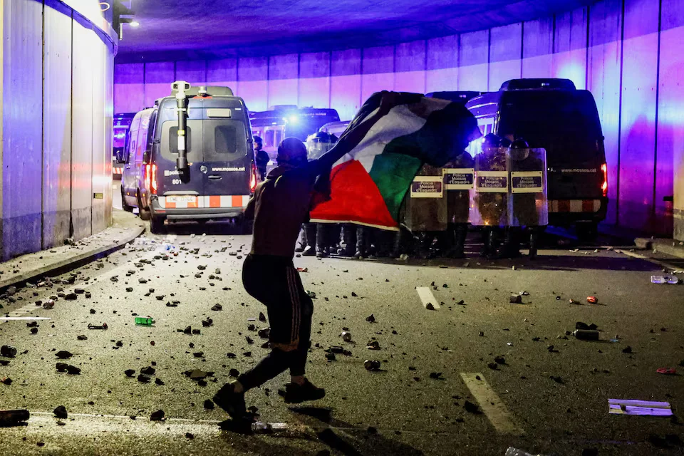 متظاهر يرفع العلم الفلسطيني أمام قوات الشرطة خلال مظاهرات أوروبية ضد اعتراض أسطول المساعدات في برشلونة (رويترز)