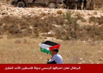 رجل يجلس على تلة رملية في فلسطين حاملًا علم فلسطين بينما تظهر قوات إسرائيلية ومركبات عسكرية في الخلفية، في سياق خبر البرتغال تعترف بدولة فلسطين